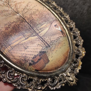 May include: A vintage oval-shaped picture frame with an ornate gold metal design. The frame holds a print of a landscape scene with trees and a figure in the distance.