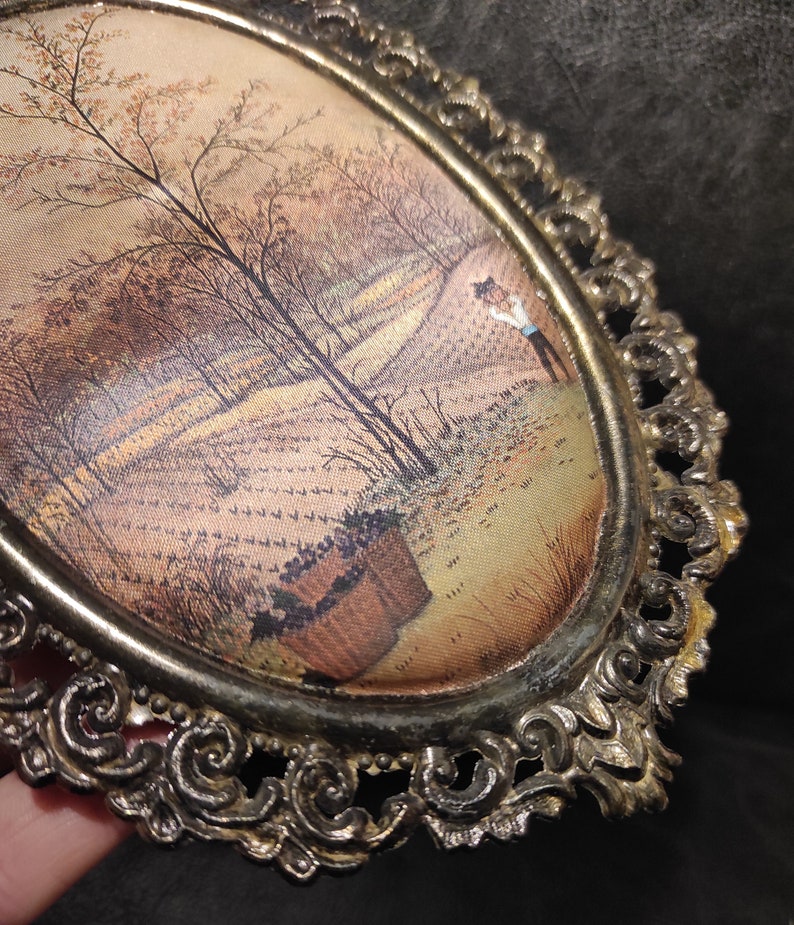 May include: A vintage oval-shaped picture frame with an ornate gold metal design. The frame holds a print of a landscape scene with trees and a figure in the distance.