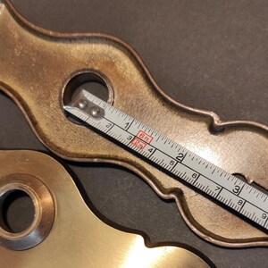 May include: Two antique brass door hardware pieces with ornate designs. A measuring tape shows a length of approximately 4 inches. The hardware has a warm, aged patina, suggesting vintage or reclaimed origins.