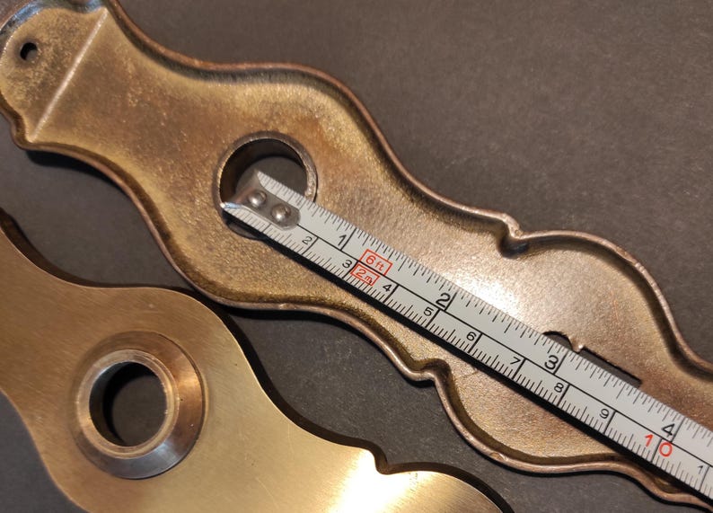 May include: Two antique brass door hardware pieces with ornate designs. A measuring tape shows a length of approximately 4 inches. The hardware has a warm, aged patina, suggesting vintage or reclaimed origins.