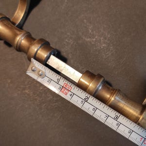 May include: A vintage brass door handle with a cylindrical design. The handle is measured with a tape measure, showing a length of approximately 4 inches. The brass has a warm, aged patina, suggesting its antique nature. The handle's design includes a series of rounded sections.