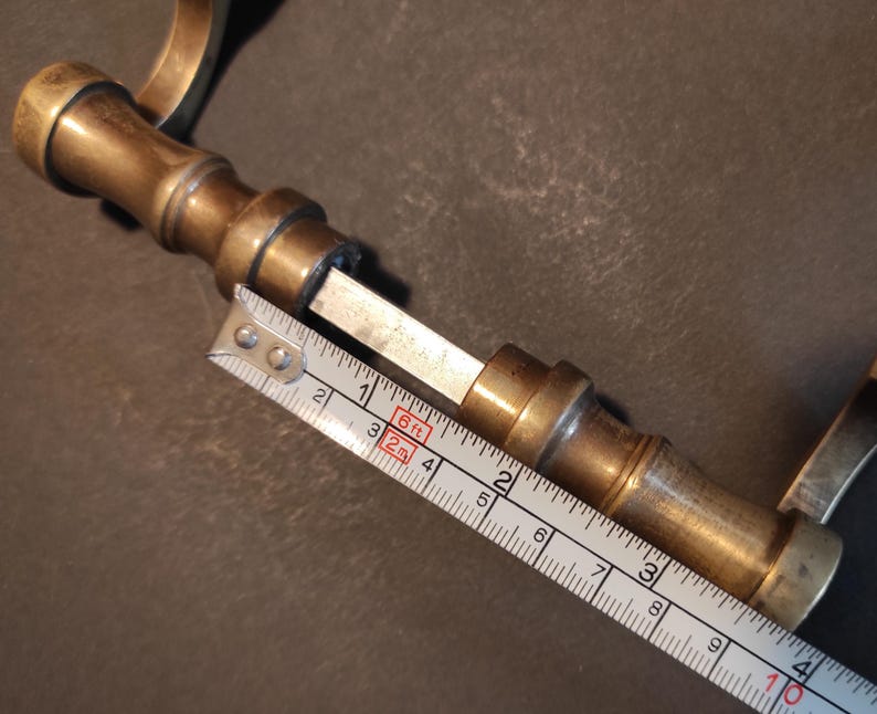 May include: A vintage brass door handle with a cylindrical design. The handle is measured with a tape measure, showing a length of approximately 4 inches. The brass has a warm, aged patina, suggesting its antique nature. The handle's design includes a series of rounded sections.