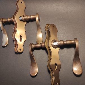 May include: Two antique brass door handles with ornate, curved designs. The handles have a warm, golden-brown hue and feature keyholes. The levers have a teardrop shape. The hardware is likely for a vintage or classic style door.