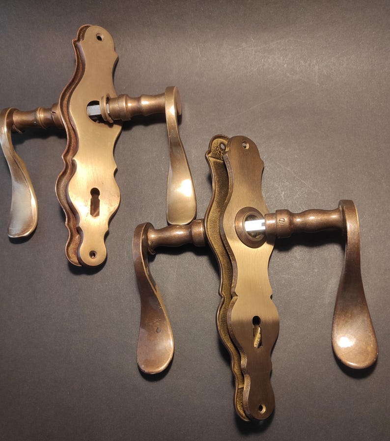 May include: Two antique brass door handles with ornate, curved designs. The handles have a warm, golden-brown hue and feature keyholes. The levers have a teardrop shape. The hardware is likely for a vintage or classic style door.