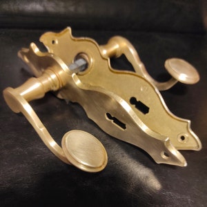 Könnte beinhalten: Ein goldfarbener Türgriff mit dekorativem Design. Der Griff hat einen Hebel und einen Knauf und ist an einer Platte mit zwei Schlüssellochern befestigt.