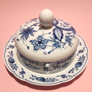 Può includere: Una burriera in ceramica bianca con un motivo floreale blu. Il piatto ha un coperchio rotondo con un pomello bianco e poggia su un piatto abbinato. Il design presenta fiori e foglie stilizzati.