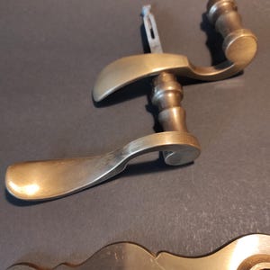 May include: Two antique brass door handles with a curved design. The handles have a warm, golden hue and a classic, vintage aesthetic. The image also shows a brass keyhole plate.