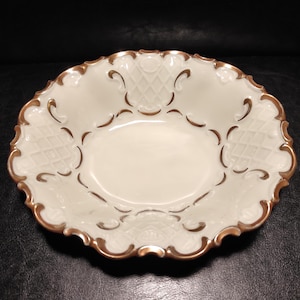 Puede incluir: Un cuenco de porcelana blanquecina con borde festoneado y ribete dorado. El cuenco presenta un centro ovalado y un diseño calado detallado alrededor del borde. El ribete dorado añade un toque de elegancia a esta fuente de servir de estilo vintage.