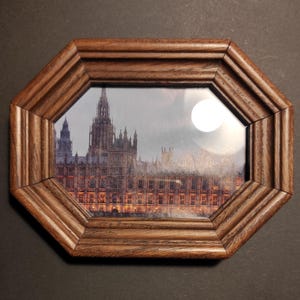Puede incluir: Un marco de fotos octogonal de madera con un borde detallado y acanalado. El marco contiene una fotografía de un edificio con múltiples torres y una fuente de luz redonda y brillante. El marco es de color marrón cálido.
