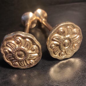 May include: Two metallic gold-toned curtain tiebacks featuring a circular, scalloped-edge face with a raised, embossed floral-inspired swirl pattern. Each tieback has a long, slender cylindrical stem extending from the back of the decorative head.