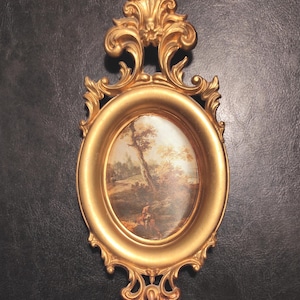 Può includere: Cornice ovale dorata e ornata con un dipinto di paesaggio all'interno. La cornice presenta un'intricata lavorazione a volute e una parte superiore decorativa. Il dipinto raffigura un albero e una figura in un ambiente naturale. La cornice è alta circa 20 cm.