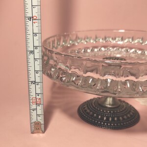 May include: A clear glass cake stand with a textured rim and a dark metal base featuring a dotted pattern. A vertical measuring tape is positioned next to the stand, showing a height of approximately 9 cm from the base to the top rim.
