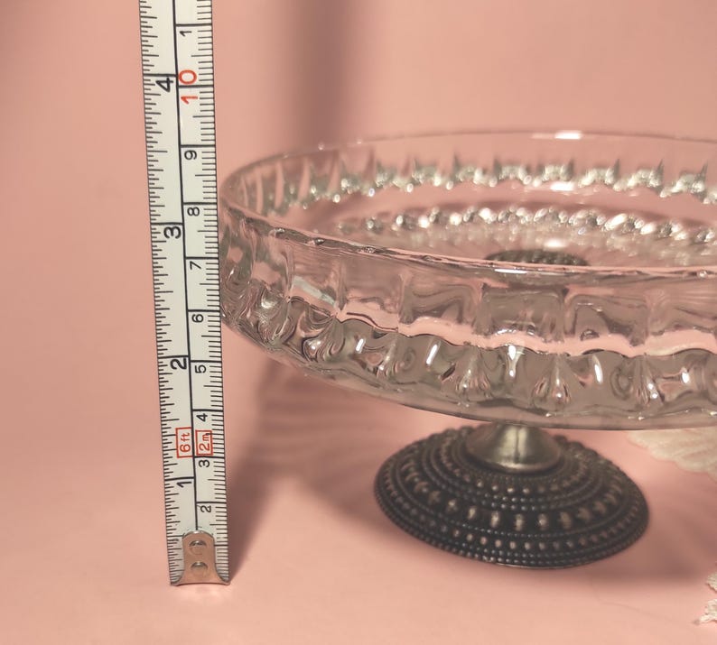 May include: A clear glass cake stand with a textured rim and a dark metal base featuring a dotted pattern. A vertical measuring tape is positioned next to the stand, showing a height of approximately 9 cm from the base to the top rim.