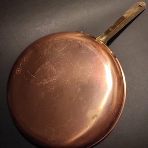 May include: A copper frying pan with a brass handle. The pan is round and has a shiny, reflective surface with visible scratches. The handle is attached with rivets and has a hole at the end for hanging. Kitchenware.