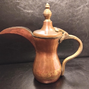 May include: A copper-colored metal coffee pot featuring a long, curved spout, a hinged lid with a pointed finial, and a textured handle. The body has a smooth, metallic finish with visible signs of wear and patina near the base and handle.