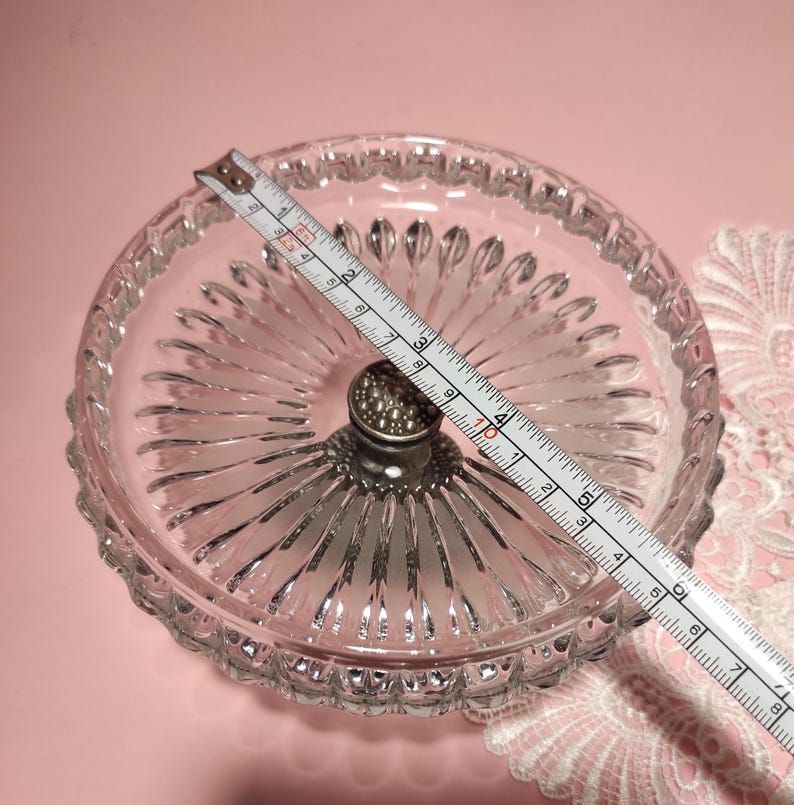 May include: A clear glass pedestal dish with a scalloped rim and a radial ribbed pattern. A silver-toned metal centre post features a textured, beaded top. A measuring tape placed across the dish shows a diameter of approximately 16.5 cm.