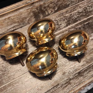 May include: Four shiny, gold-colored cabinet knobs with a rounded, oval shape. Each knob has a threaded post for mounting. The knobs are arranged on a weathered, wooden surface, creating a vintage aesthetic.