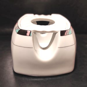 May include: A white ceramic creamer with a spout and a circular opening at the top. The creamer has a geometric design in pink, black, and teal. A thin black line accents the base of the creamer.