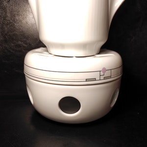 Puede incluir: Tetera de cerámica blanca con un calentador de cerámica blanca a juego. El calentador tiene un diseño circular con una línea negra y un pequeño círculo rosa.