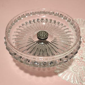 May include: A clear glass dish featuring a radial sunburst pattern on the base and a textured rim. A small, circular silver-toned metal knob with a raised, dotted surface is positioned in the centre of the dish, serving as a decorative handle.