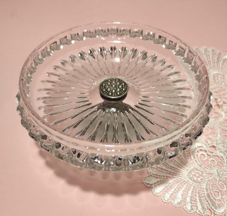 May include: A clear glass dish featuring a radial sunburst pattern on the base and a textured rim. A small, circular silver-toned metal knob with a raised, dotted surface is positioned in the centre of the dish, serving as a decorative handle.