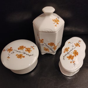 Könnte beinhalten: Drei weiße Porzellanboxen mit Blumendesigns. Die Boxen sind mit einem zarten Zweig mit orangefarbenen Blüten verziert. Die Boxen haben alle unterschiedliche Formen und Größen.