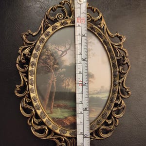 Puede incluir: Un marco de fotos ovalado de lat&oacute;n antiguo con un dise&ntilde;o ornamental y ondulado. El marco contiene una pintura de paisaje. El marco mide aproximadamente 15 cm de altura. El fondo es de color oscuro y neutro.