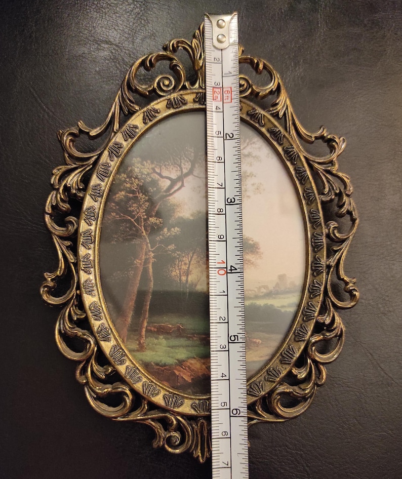 Puede incluir: Un marco de fotos ovalado de lat&oacute;n antiguo con un dise&ntilde;o ornamental y ondulado. El marco contiene una pintura de paisaje. El marco mide aproximadamente 15 cm de altura. El fondo es de color oscuro y neutro.