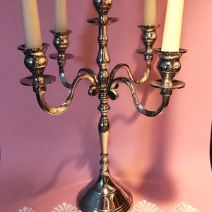 May include: A silver-colored candelabra with five arms, each holding a cream-colored candle. The candelabra has a decorative, curved design and sits on a round base. The background is a solid pink color.