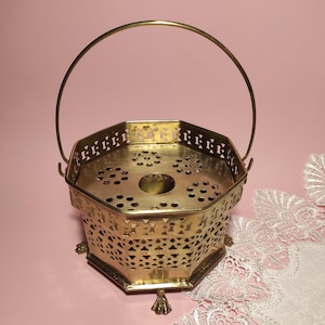 Könnte beinhalten: Ein dekorativer Vintage-Korb aus Messing mit Griff. Der achteckige Korb hat ein perforiertes Design an den Seiten und ein kreisförmiges Muster auf der Oberseite. Der Korb wird von drei kleinen Füßen getragen und steht auf einer rosa Oberfläche.