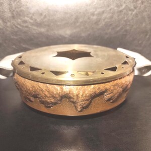 May include: A round, two-toned decorative container with a textured, bronze-coloured base and a silver-coloured lid. The lid has a star-shaped cutout and a decorative brass rim. White fabric handles are attached to the sides.