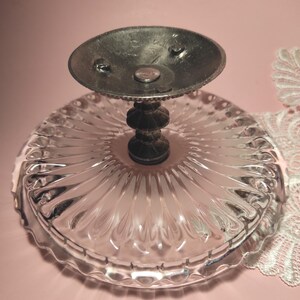 May include: A clear glass pedestal dish featuring a circular plate with a radial ribbed pattern and a scalloped edge. The base is supported by a dark metallic stem with a tiered design and a circular metal plate at the very top of the stand.