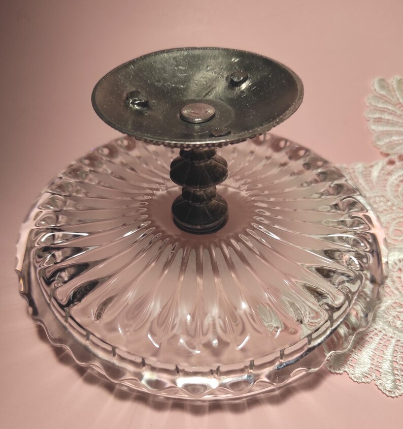 May include: A clear glass pedestal dish featuring a circular plate with a radial ribbed pattern and a scalloped edge. The base is supported by a dark metallic stem with a tiered design and a circular metal plate at the very top of the stand.