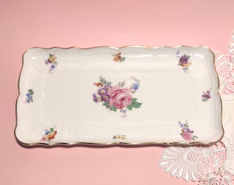 May include: A rectangular white porcelain serving tray with scalloped edges and a gold-toned rim. The center features a floral bouquet with a large pink rose, while smaller floral clusters are distributed around the border on a white surface.