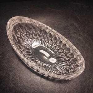 May include: Clear, oval-shaped crystal dish with a textured, leaf-like pattern. The dish is empty and reflects light, showcasing its intricate design. The crystal dish is a decorative item.