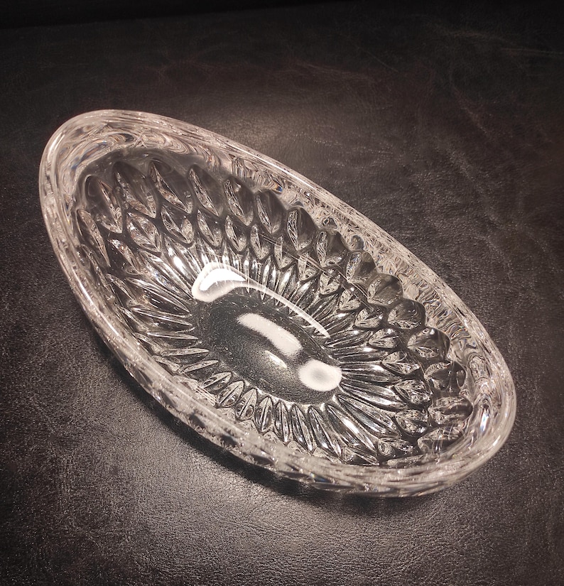 May include: Clear, oval-shaped crystal dish with a textured, leaf-like pattern. The dish is empty and reflects light, showcasing its intricate design. The crystal dish is a decorative item.