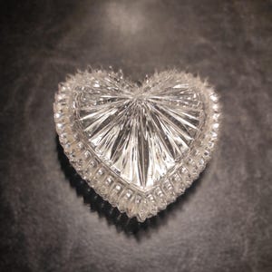 May include: A clear, heart-shaped trinket box with a textured, cut-glass design. The box features a detailed, ridged pattern radiating from the center and a scalloped edge. The box is displayed on a dark surface.