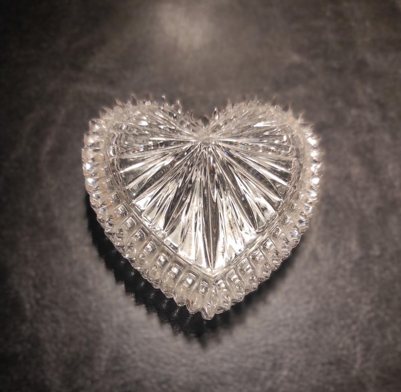May include: A clear, heart-shaped trinket box with a textured, cut-glass design. The box features a detailed, ridged pattern radiating from the center and a scalloped edge. The box is displayed on a dark surface.