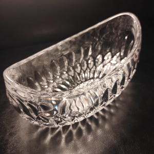 May include: A clear, oval-shaped glass bowl with a textured, cut-glass design. The bowl's surface reflects light, creating a sparkling effect. The bowl is likely used for serving or display.