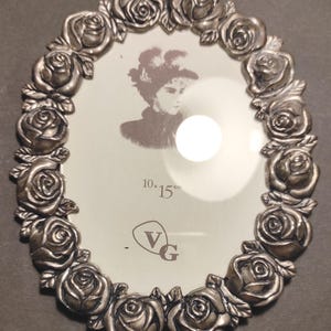 Peut inclure: Cadre photo ovale argenté orné de roses. Il contient une image en noir et blanc et porte la mention "10 x 15 cm". Ce cadre décoratif est idéal pour afficher des photos.