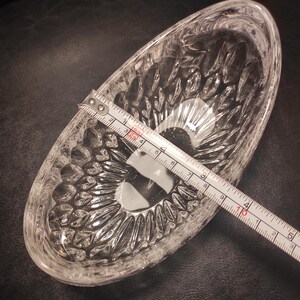 May include: A clear, oval-shaped glass dish with a textured, cut-glass design. A measuring tape shows the dish is approximately 5 inches long. The dish is empty and sits on a dark surface.