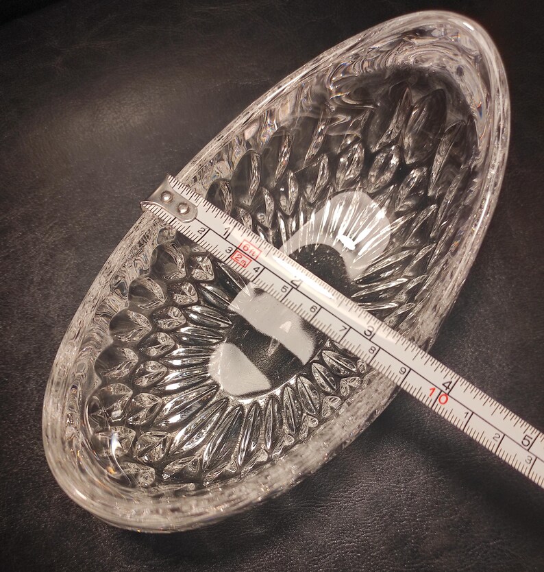 May include: A clear, oval-shaped glass dish with a textured, cut-glass design. A measuring tape shows the dish is approximately 5 inches long. The dish is empty and sits on a dark surface.