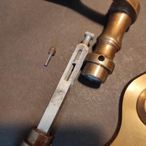 May include: Close-up of disassembled antique brass door hardware. The components include a handle, a long metal piece with a screw, and a small screw. The hardware has a tarnished, aged appearance.