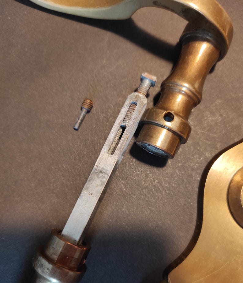 May include: Close-up of disassembled antique brass door hardware. The components include a handle, a long metal piece with a screw, and a small screw. The hardware has a tarnished, aged appearance.