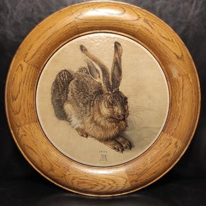 May include: A framed print of a brown hare, with long ears and detailed fur, resting on a light background. The print is set within a round, wooden frame. The year 1502 is visible in the lower left corner of the print.