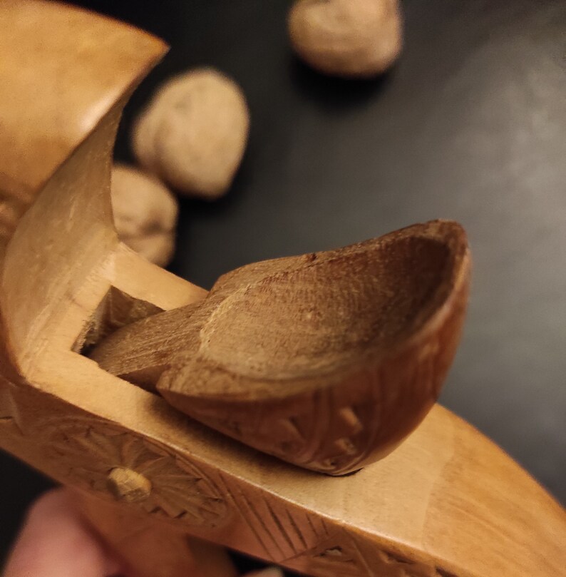 Op de afbeelding: Een houten notenkraker met een gesneden ontwerp. De notenkraker heeft een scharnierende kaak en een kleine, gesneden schep om noten vast te houden.