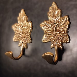 Puede incluir: Dos ganchos de metal dorado en forma de hojas estilizadas con detalles intrincados. Los ganchos están diseñados para colgar objetos en una pared.