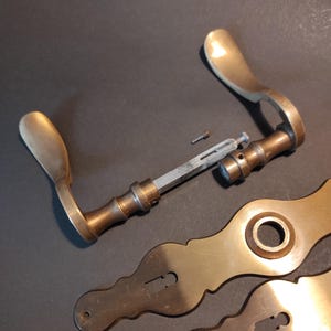 May include: Antique brass door hardware set, including a handle, escutcheons, and connecting components. The handle has a curved design with a warm, golden hue. The escutcheons feature decorative shapes. The hardware is set against a dark background.