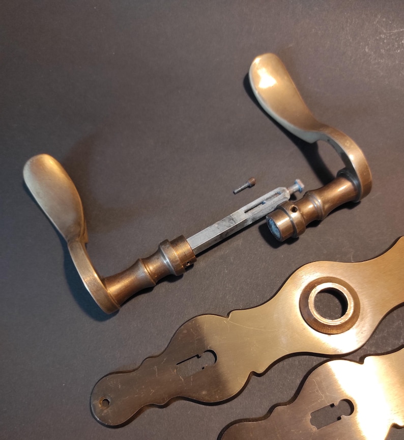 May include: Antique brass door hardware set, including a handle, escutcheons, and connecting components. The handle has a curved design with a warm, golden hue. The escutcheons feature decorative shapes. The hardware is set against a dark background.