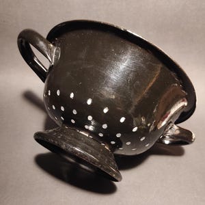 May include: A black metal colander with two handles and a pedestal base. The colander features small, evenly spaced white dots. The kitchen utensil has a glossy finish.